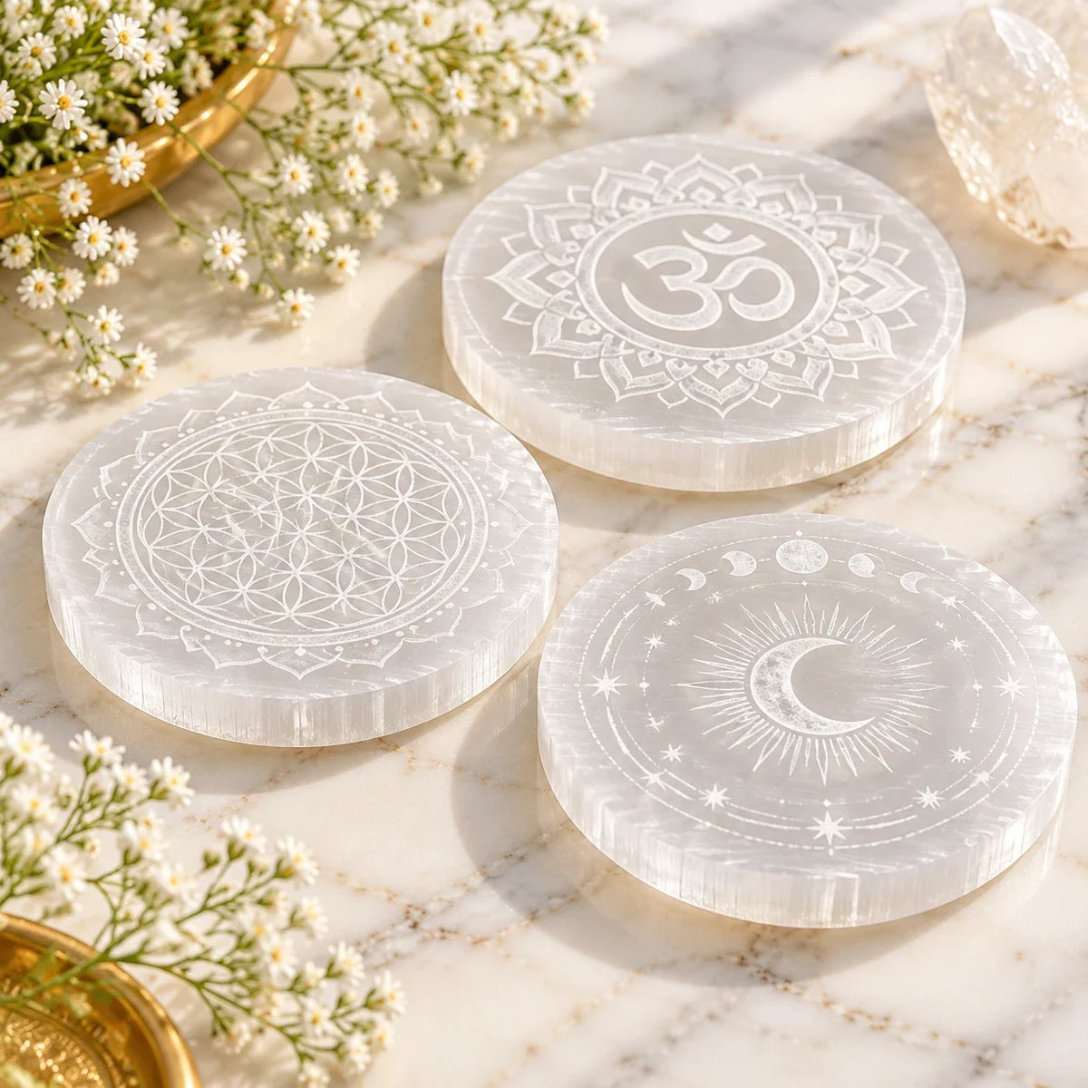 Three engraved selenite charging plates with Om, Flower of Life and crescent moon designs on white marble